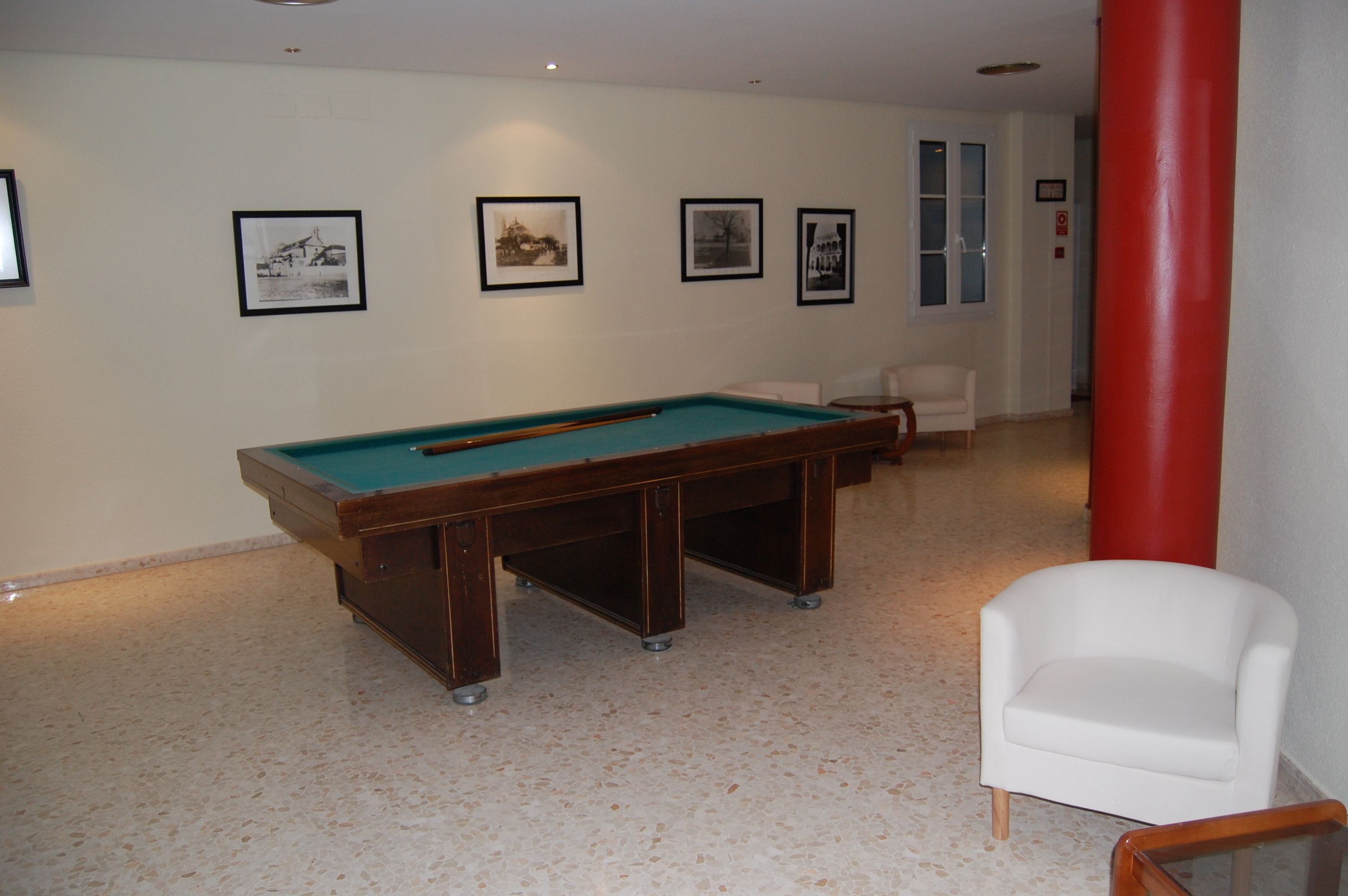 Une table de billard dans une pièce dont les murs sont ornés de tableaux encadrés et où se trouve une chaise blanche.