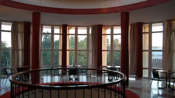 A spacious interior with a central circular railing, large windows offering a view of greenery, and multiple tables and chairs arranged around the space.