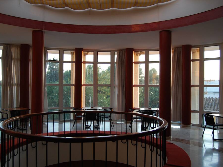 A spacious interior with a central circular railing, large windows offering a view of greenery, and multiple tables and chairs arranged around the space.