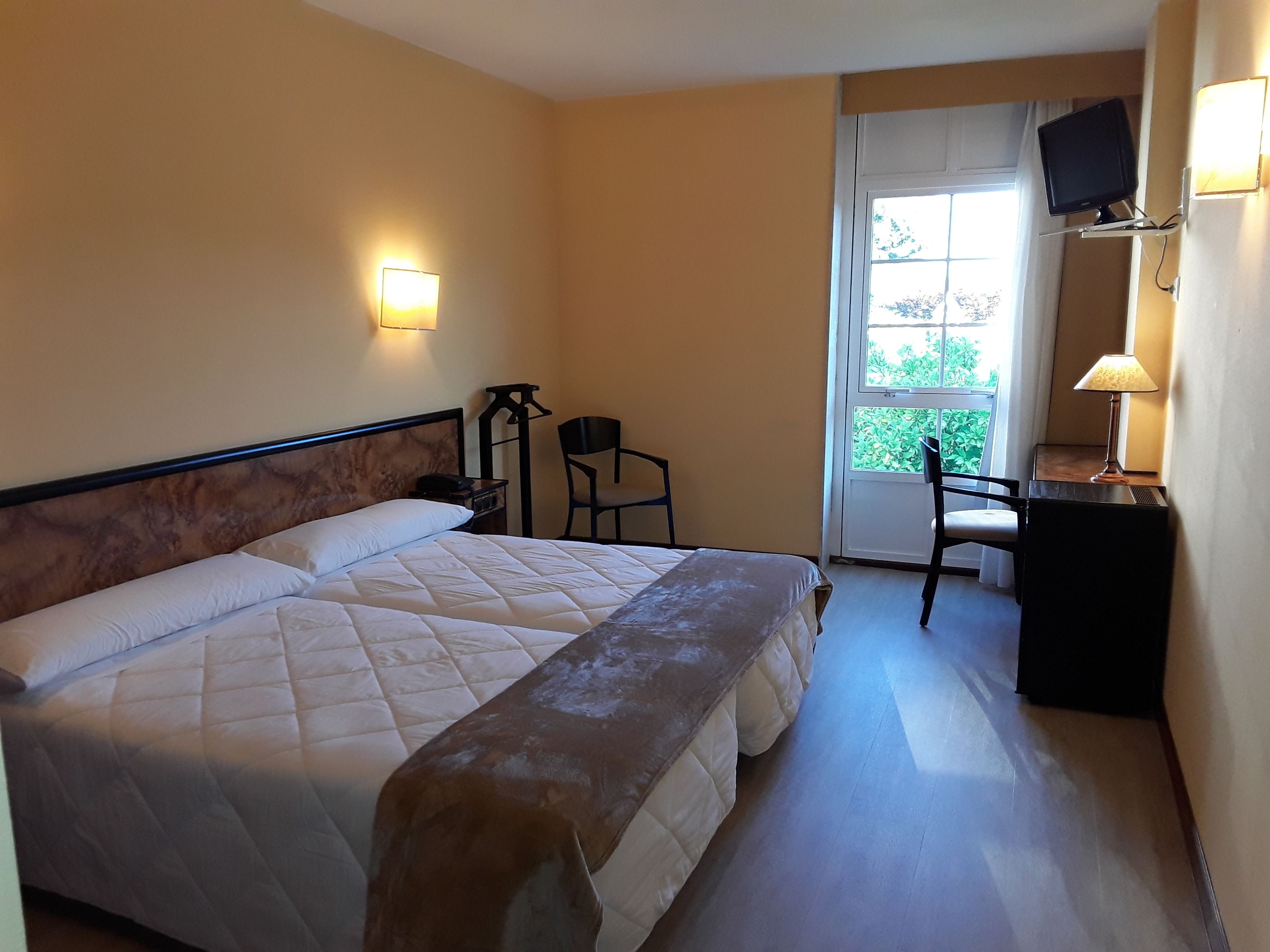 A hotel room with a large bed, a desk with a chair, a television mounted on the wall, and a window with a view of greenery.