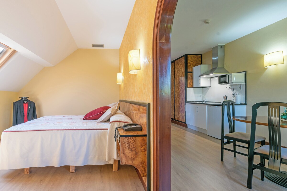 A bedroom with a bed, bedside table, and a telephone. There is a window providing natural light and a view of the outdoors.