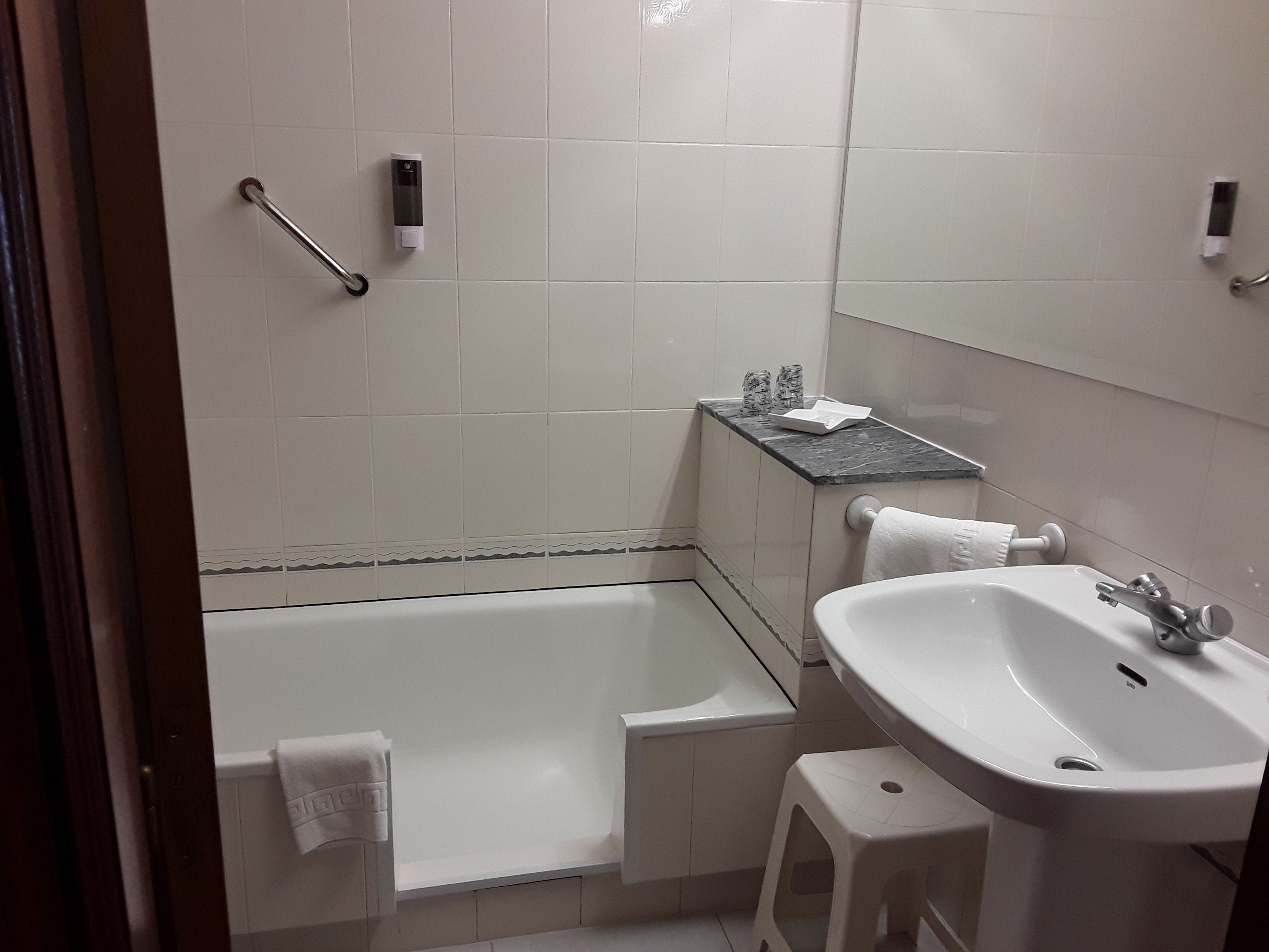 Um banheiro com banheira, pia e um pequeno banquinho. Há uma toalha pendurada na parede e um dosador de sabonete no balcão.