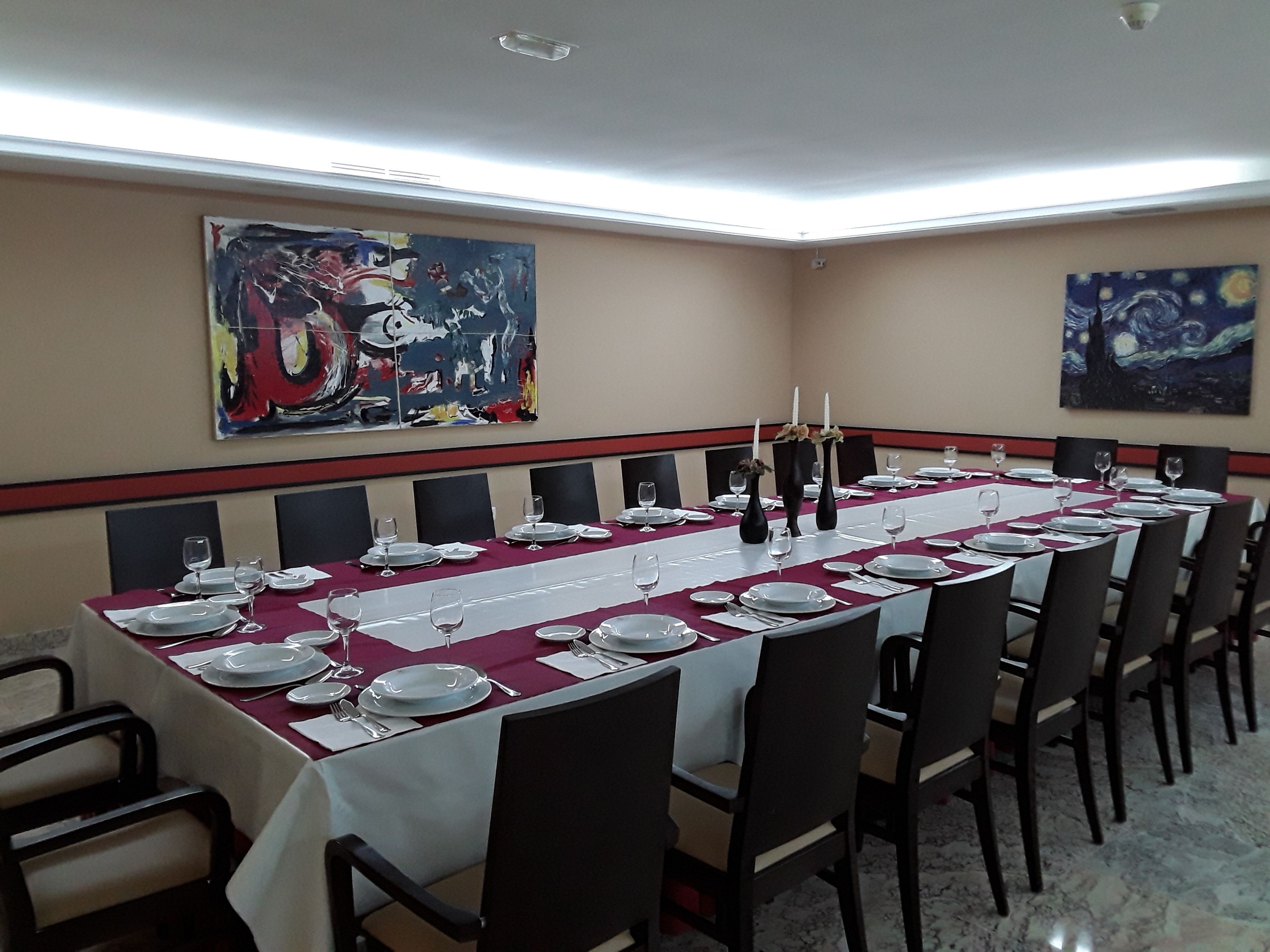 A conference room with a long table set for a meeting, featuring a white tablecloth, black chairs, and two paintings on the walls.
