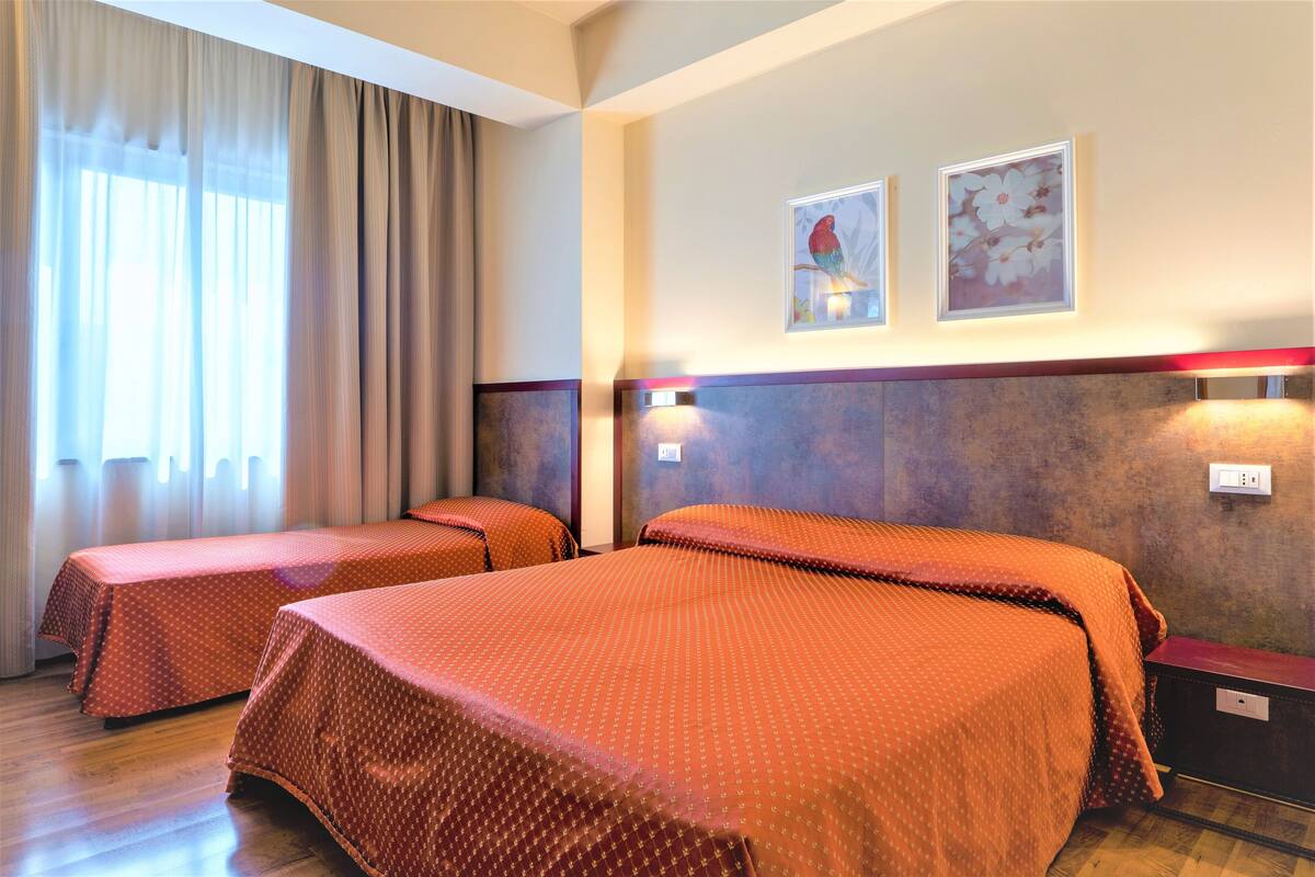 A hotel room with two beds, a large window with curtains, and two framed pictures on the wall.