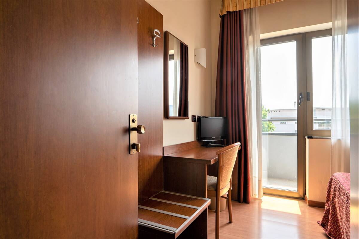 A hotel room with a wooden desk, a chair, a television, and a balcony with curtains.