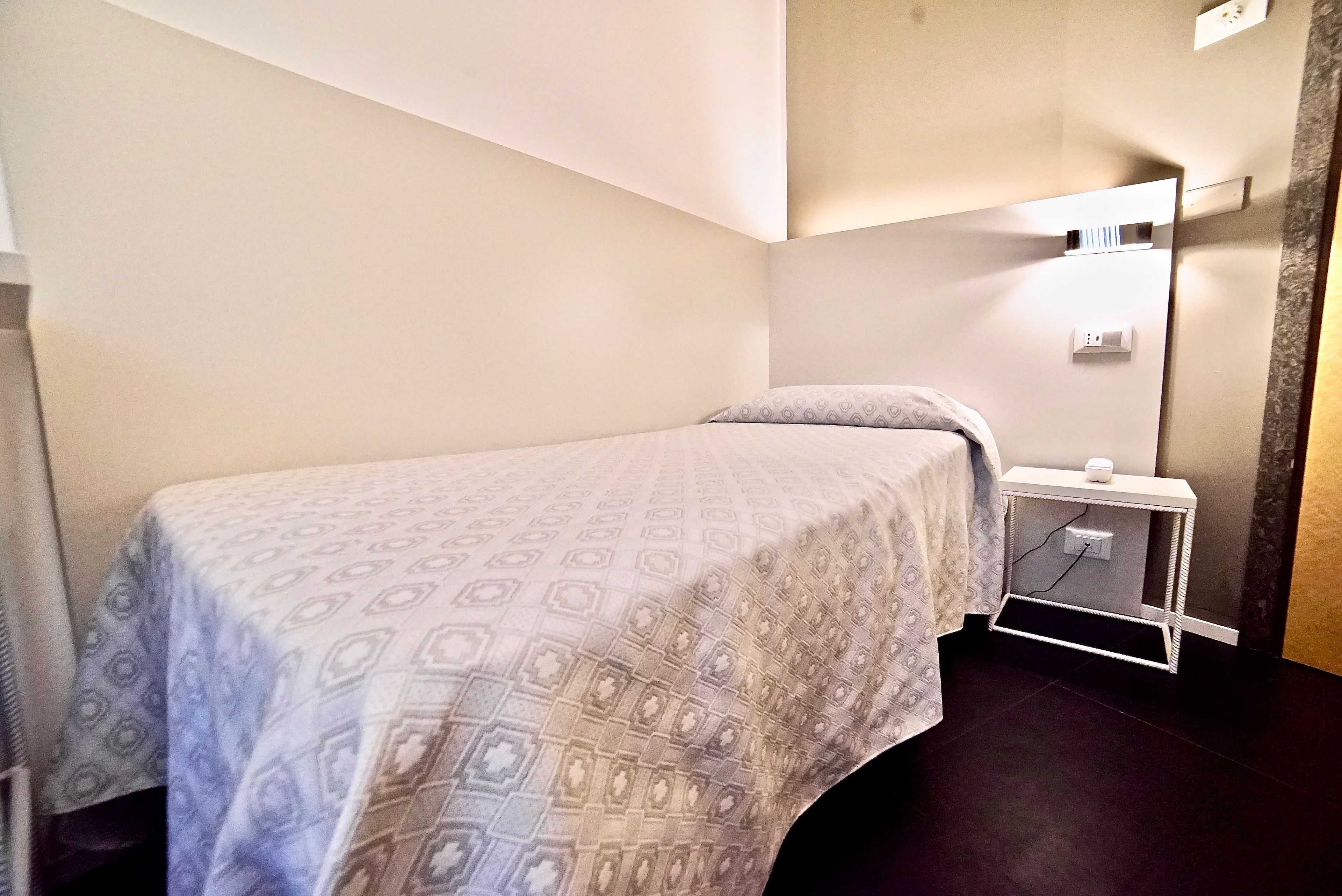 Een klein, net bed met een gestreept beddekleed in een kamer met een hoekvenster, een klein nachtkastje en een wandlamp.