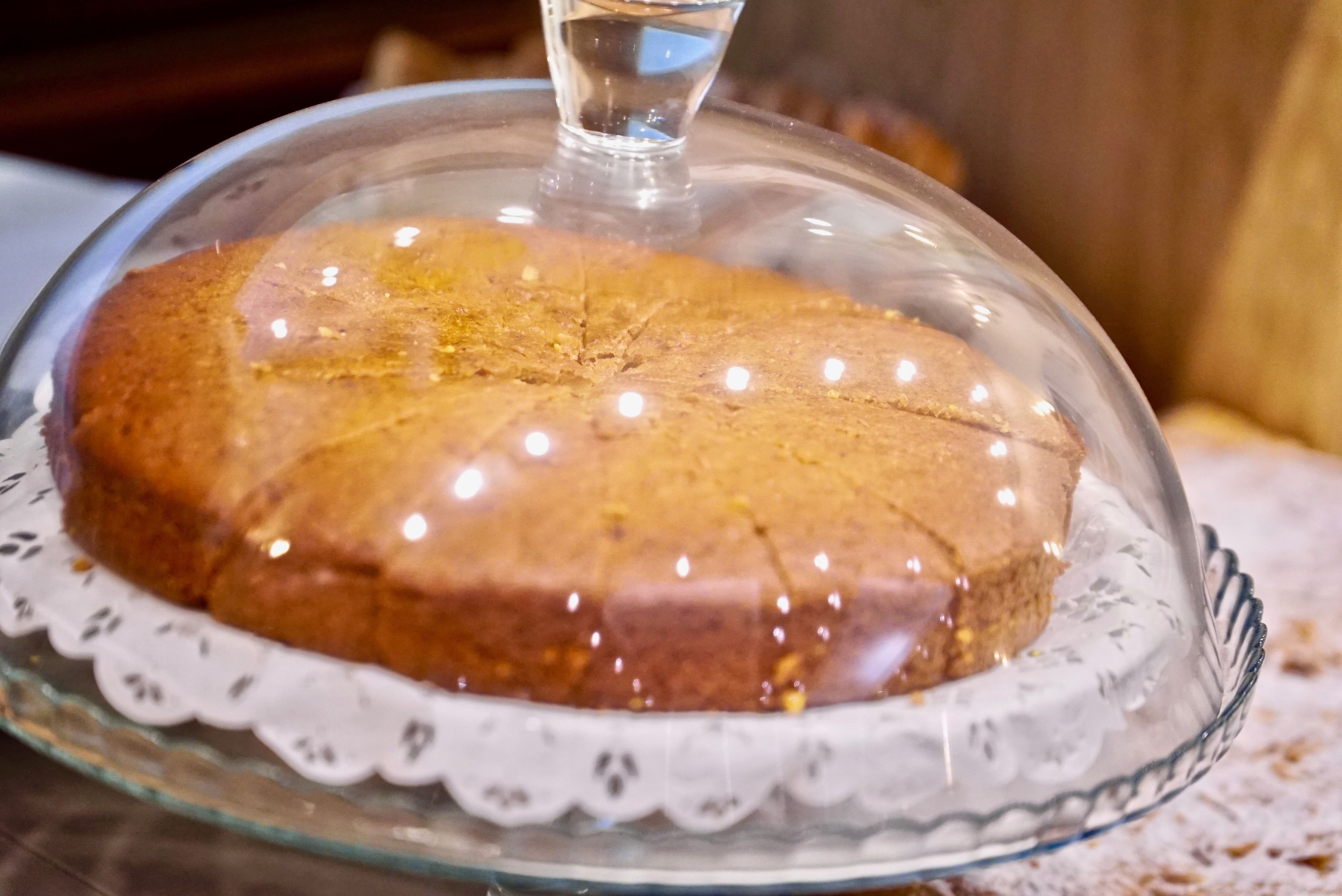 Un pastel redondo con una campana de cristal sobre un plato de cristal, colocado sobre un mantel de encaje.