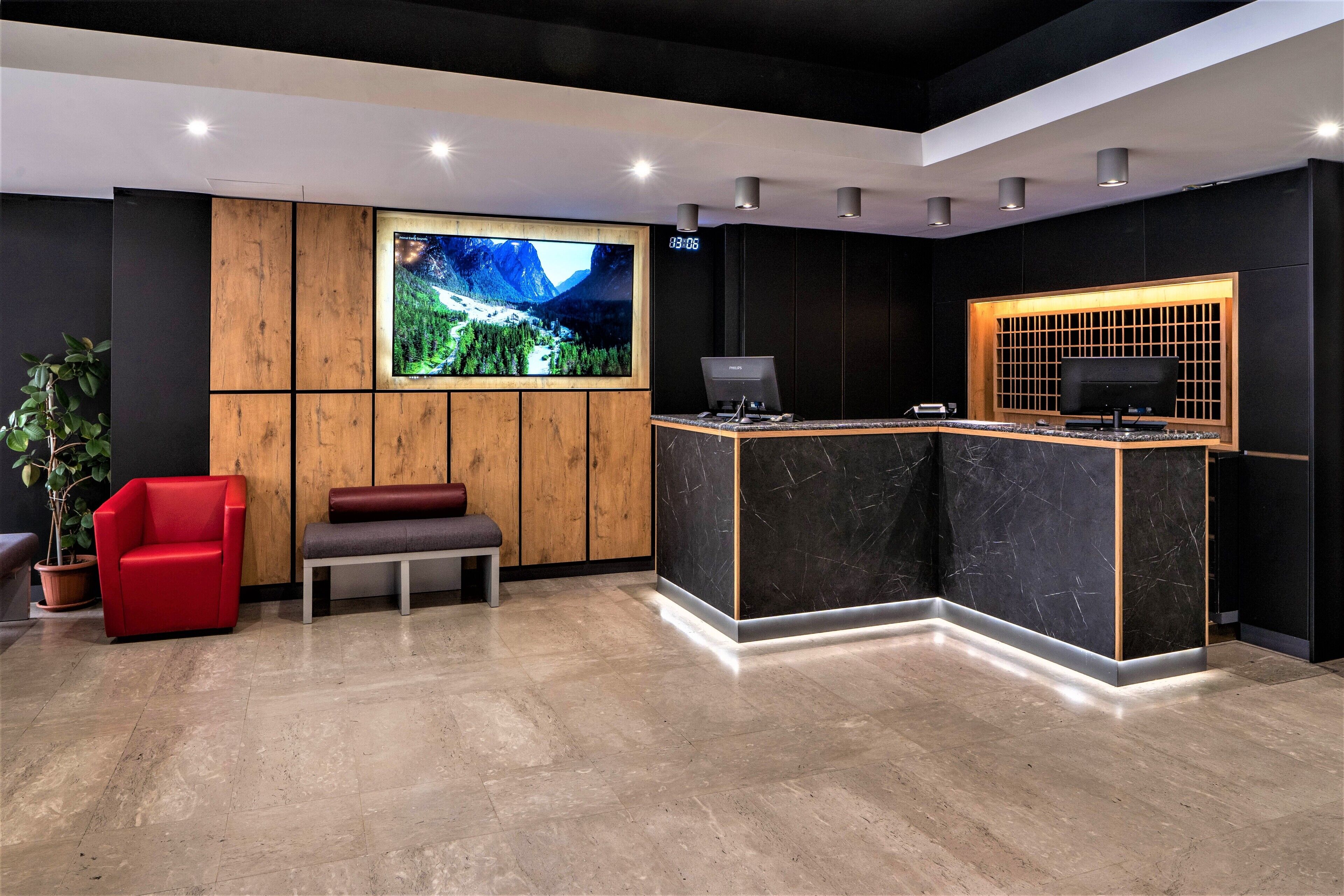 A reception area with a wooden panel wall, a large flat-screen TV, a reception desk with a computer, a red chair, and a bench.