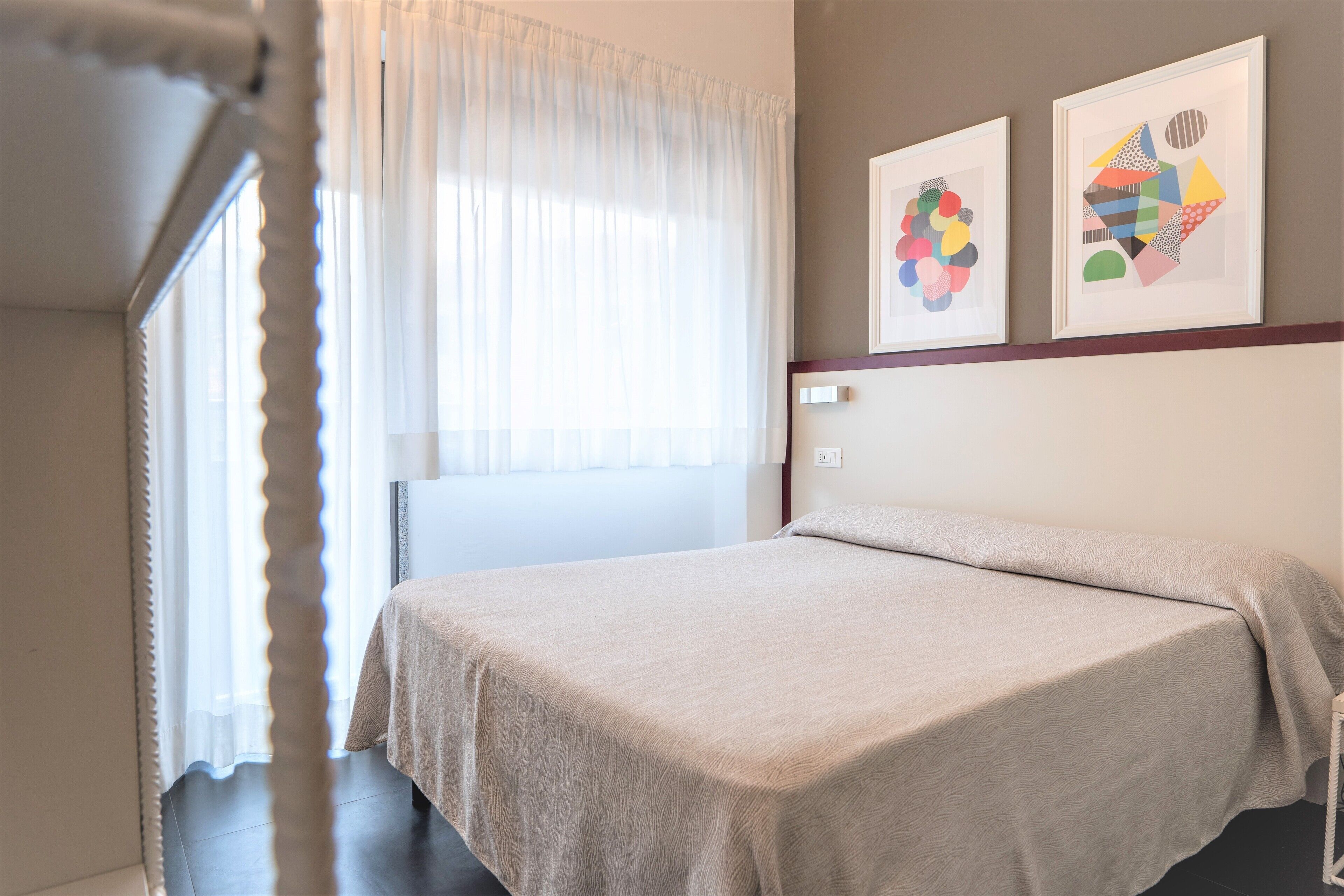 A bedroom with a bed, a window with sheer curtains, and two framed abstract artworks on the wall.