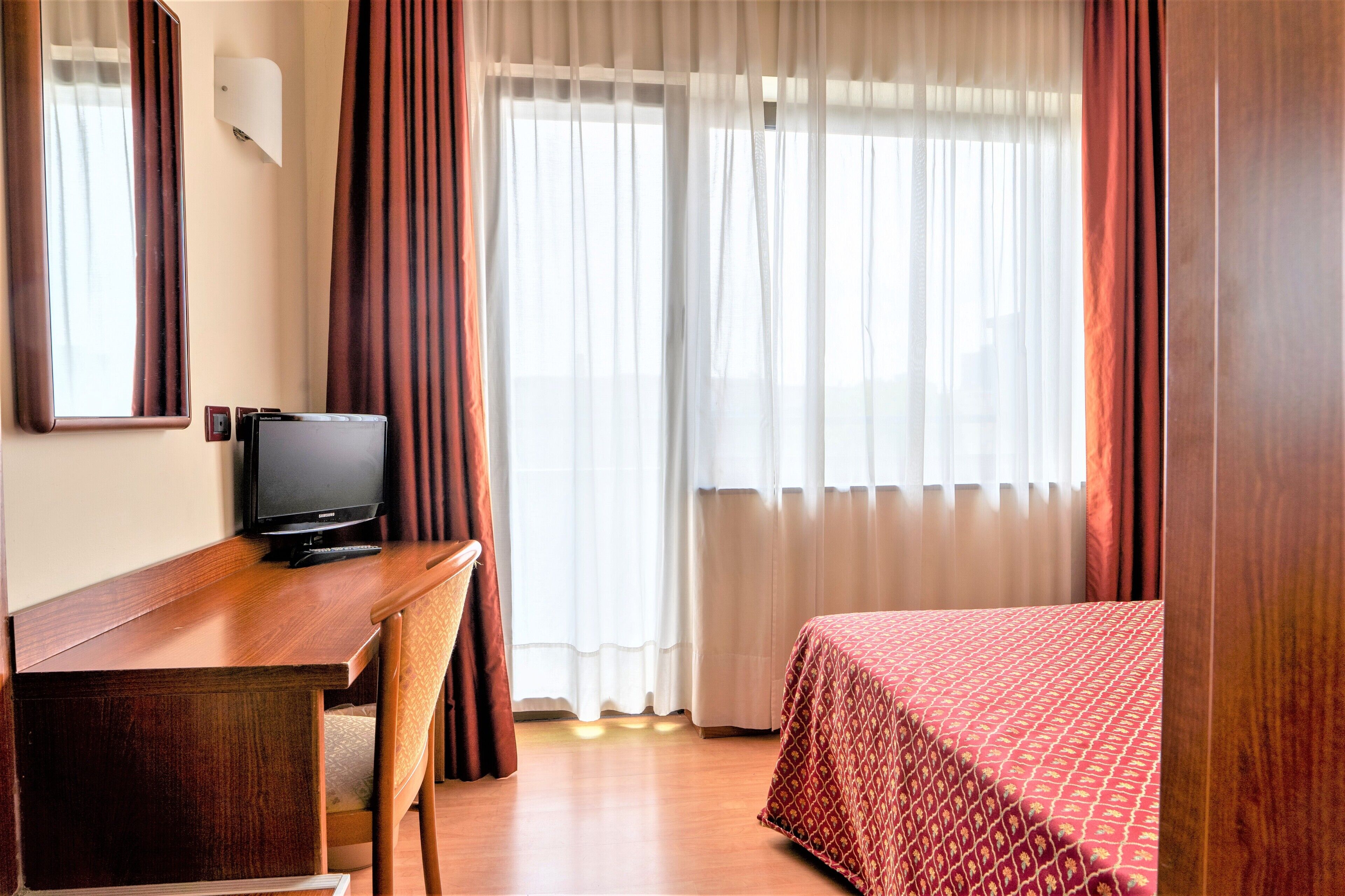 A hotel room with a wooden desk, a television, a bed with a patterned bedspread, and a window with sheer curtains.