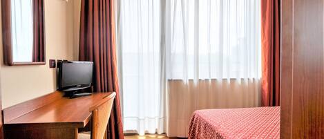 A hotel room with a wooden desk, a television, a bed with a patterned bedspread, and a window with sheer curtains.