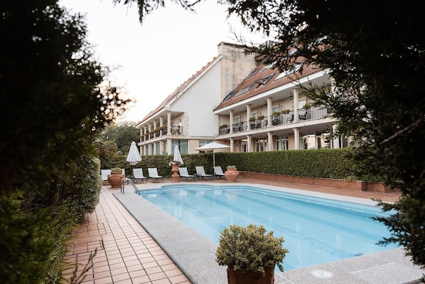 Un área junto a la piscina con sillones de descanso y sombrillas, un edificio con varios balcones y un seto al frente.