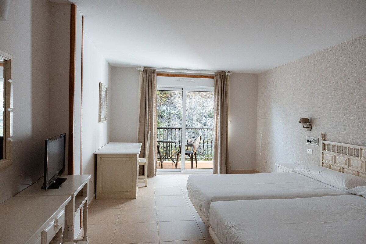 Habitación de hotel con cama, escritorio, televisor, balcón con mesa y sillas, y ventana con cortinas.
