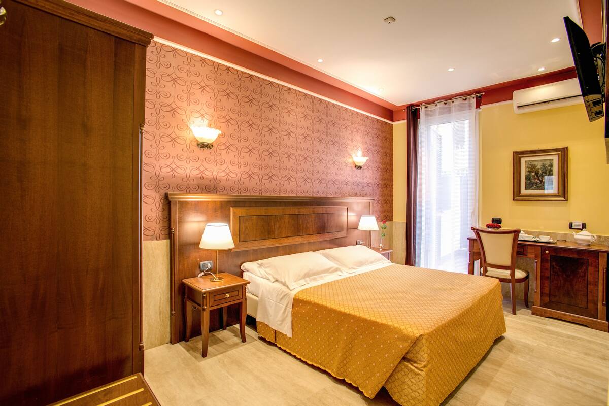 A hotel room with a wooden headboard, two bedside lamps, a desk with a chair, and a large bed with a yellow bedspread.