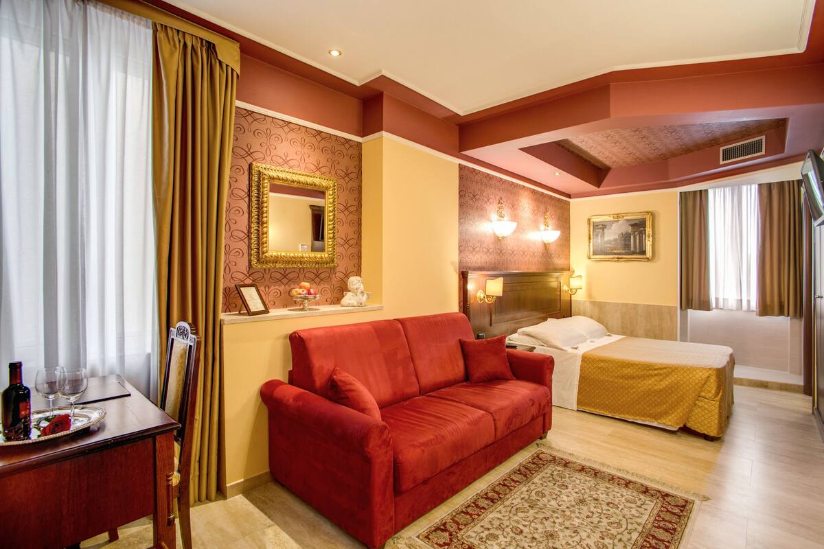 A hotel room with a red sofa, a bed with a yellow bedspread, a wooden nightstand, a mirror, and a wall-mounted light fixture.