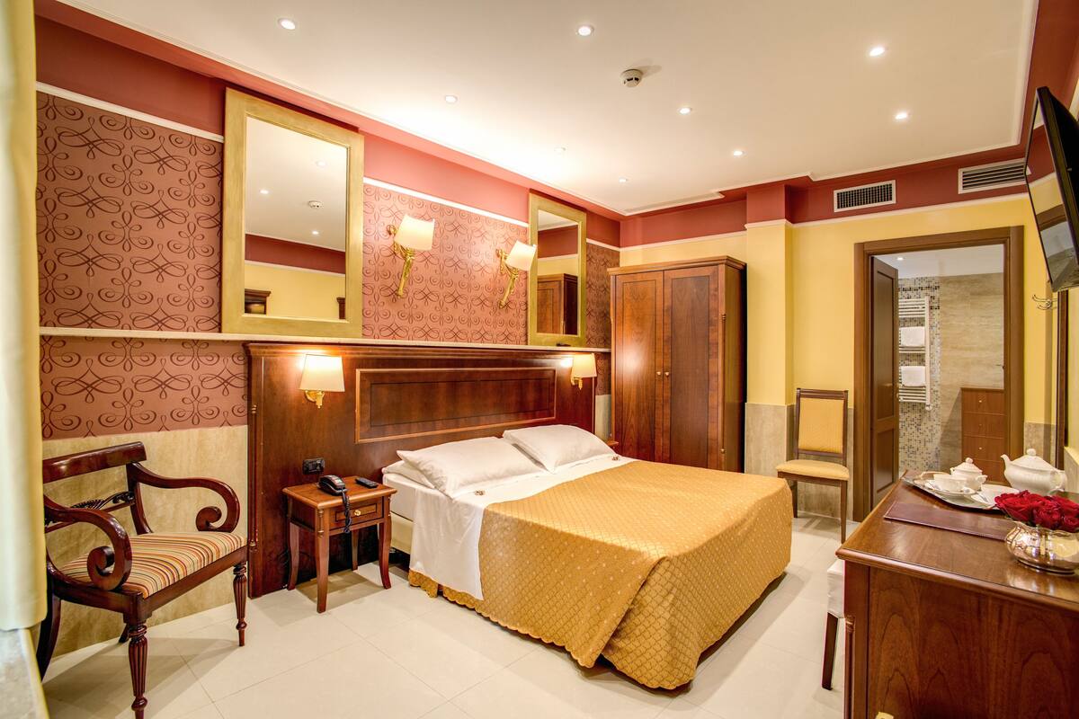 A hotel room with a large bed, a wooden chair, a desk with a telephone, and a mirror on the wall.