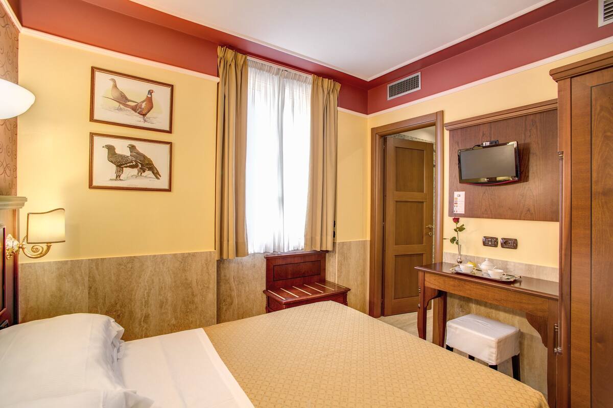 A hotel room with a bed, a desk with a television, a small bench, and two framed pictures on the wall.
