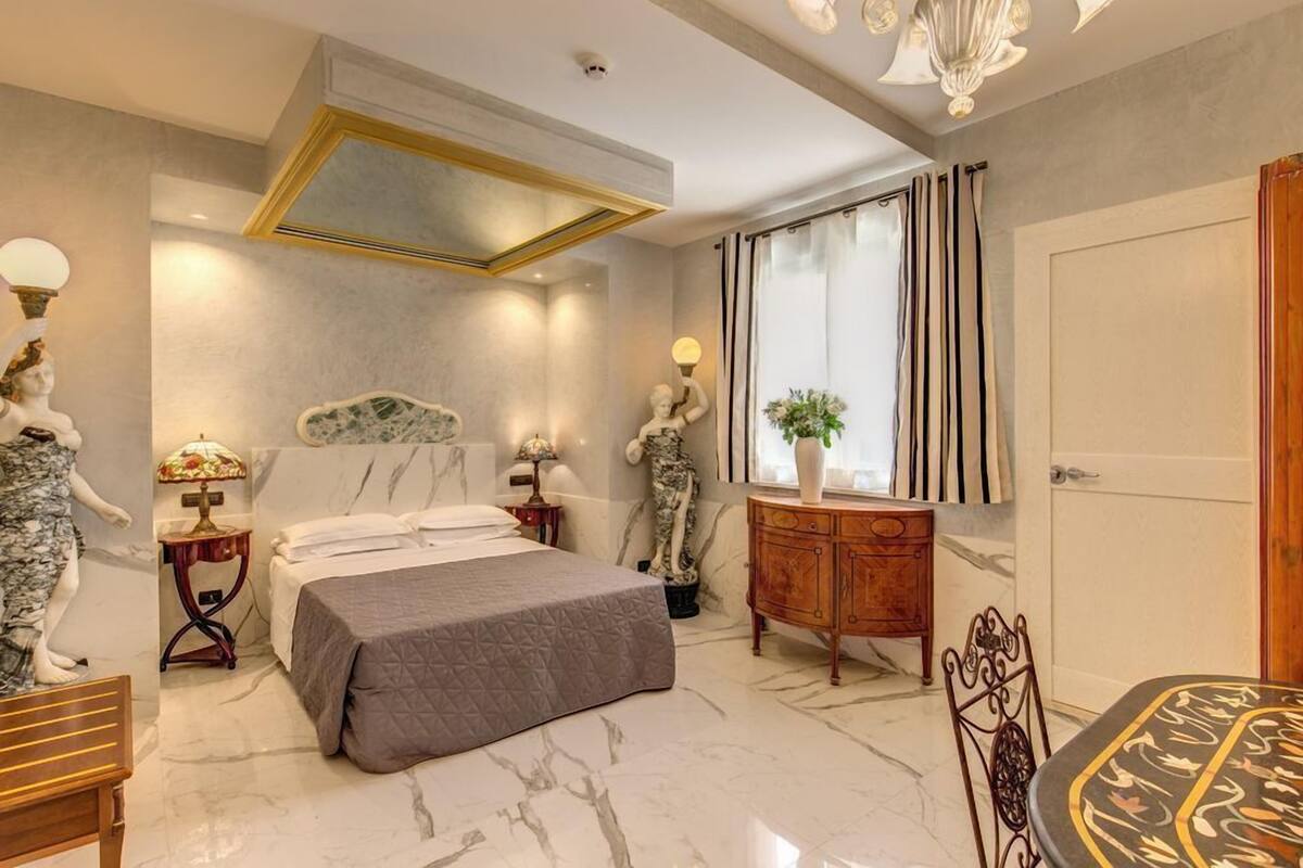 A modern bedroom with a marble floor, a bed with a grey quilt, a wooden dresser, and a dining table with a patterned tablecloth.