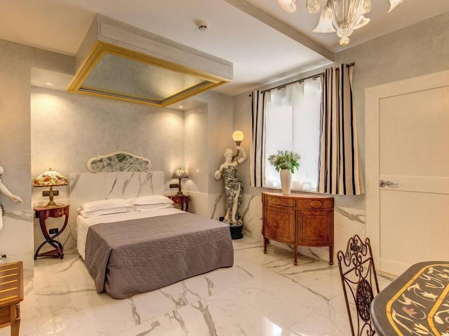 A modern bedroom with a marble floor, a bed with a grey quilt, a wooden dresser, and a dining table with a patterned tablecloth.