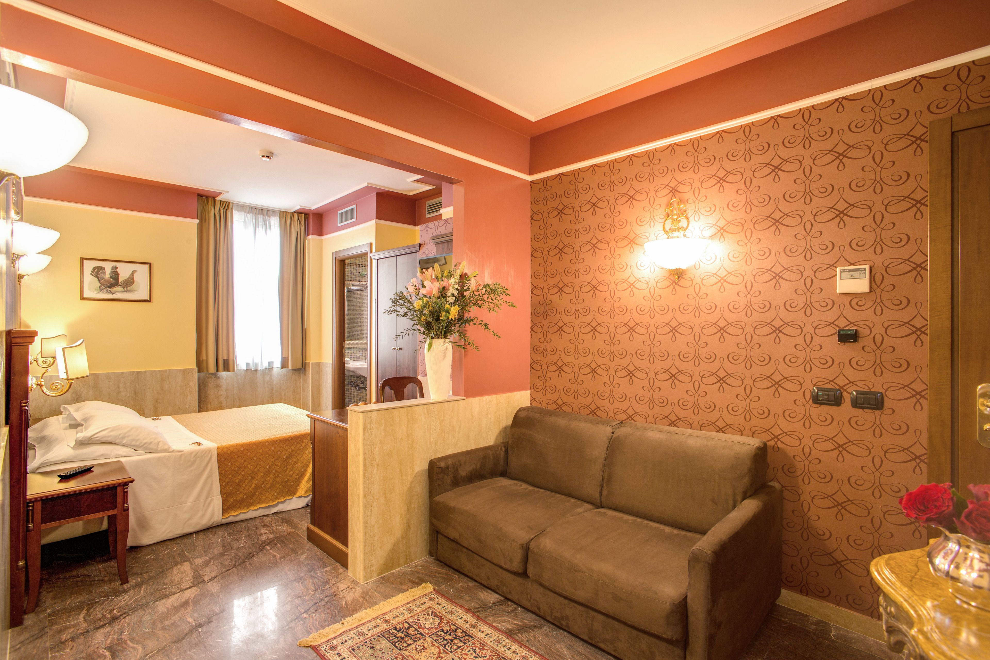 A hotel room with a bed, a sofa, a desk, a lamp, a vase with flowers, and a patterned wall.