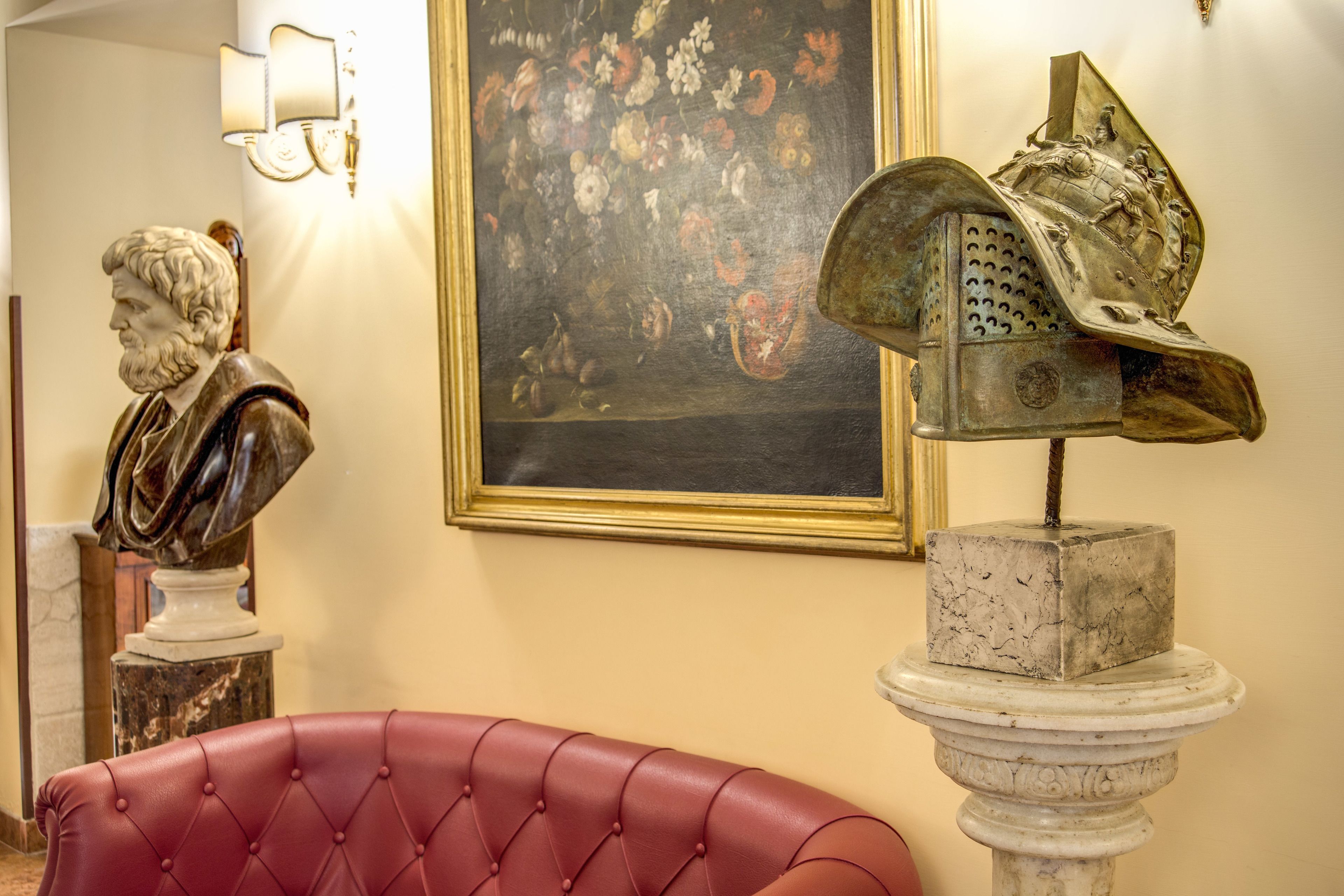 Une pièce comprenant un buste en marbre, un tableau floral encadré et un fauteuil recouvert d’un tissu rouge.