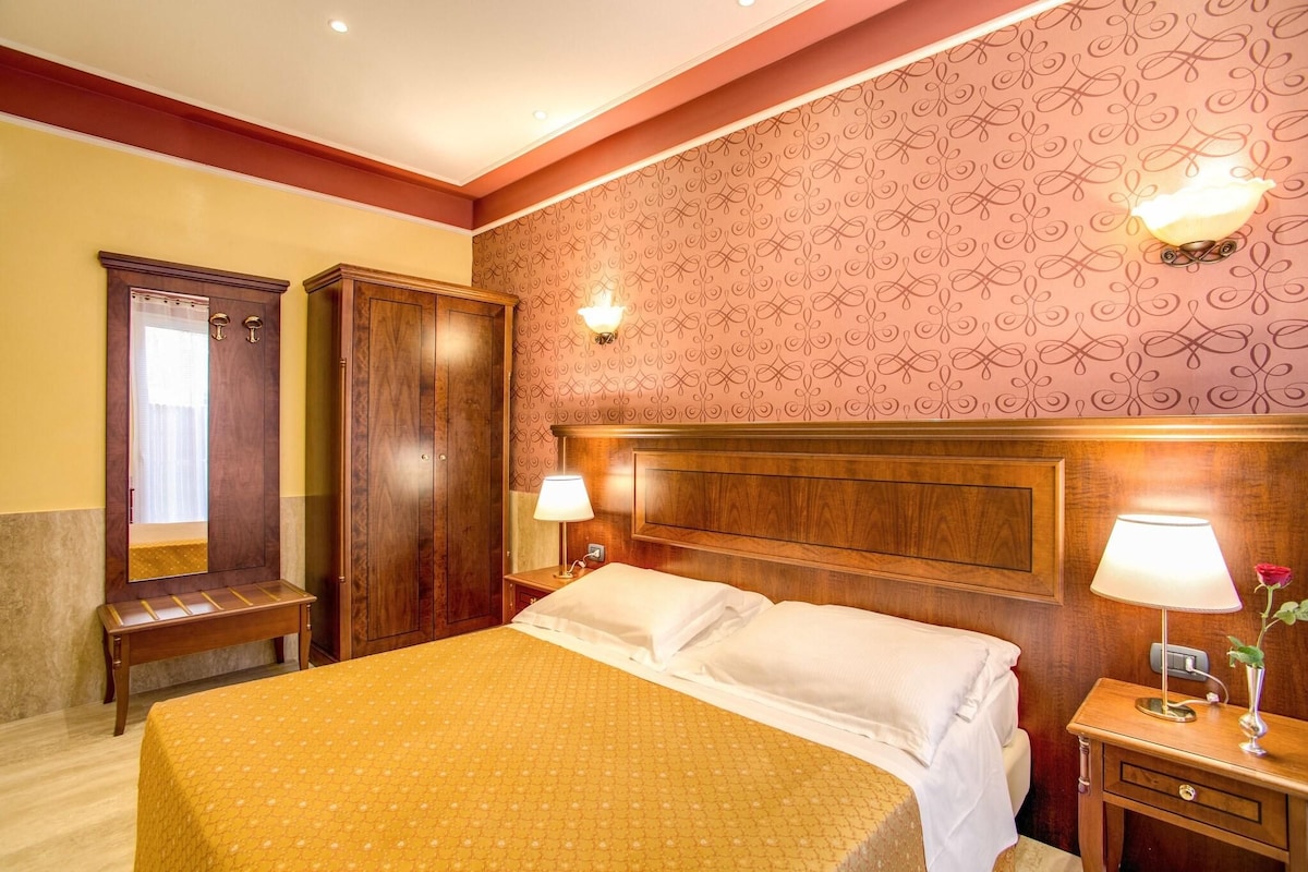 A hotel room with a wooden headboard, two bedside tables with lamps, a bench, and a large bed with a patterned bedspread.