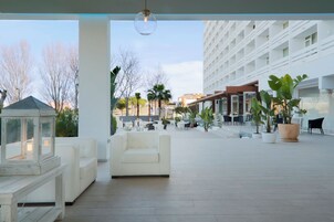 Amplia zona de estar al aire libre con mobiliario blanco, plantas en macetas y vista despejada de un edificio moderno al fondo.