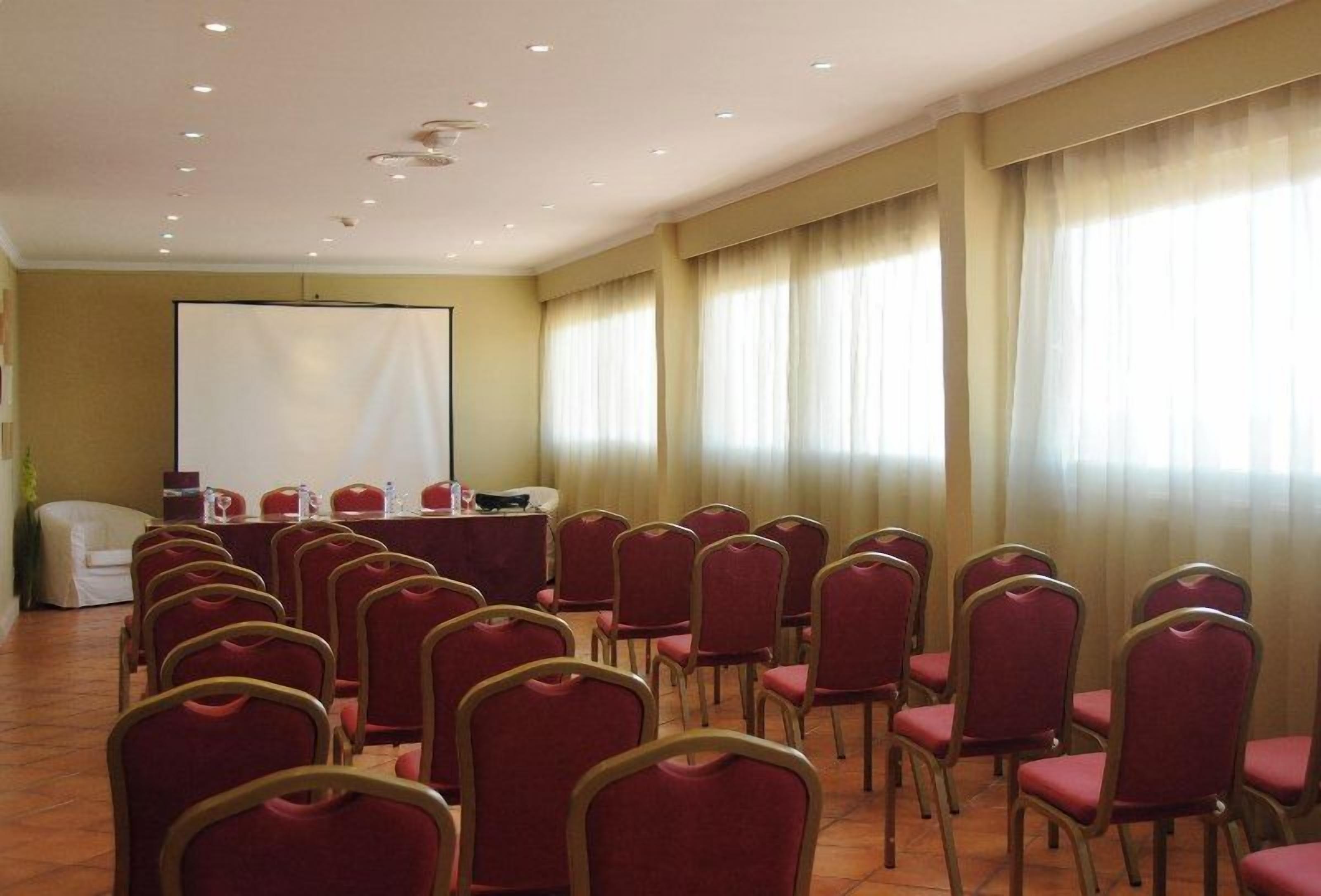 A conference room with rows of red chairs, a white table with microphones, and a large screen at the front.