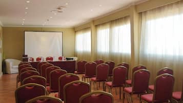 A conference room with rows of red chairs, a white table with microphones, and a large screen at the front.