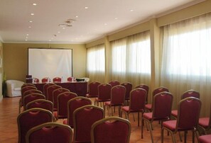 A conference room with rows of red chairs, a white table with microphones, and a large screen at the front.