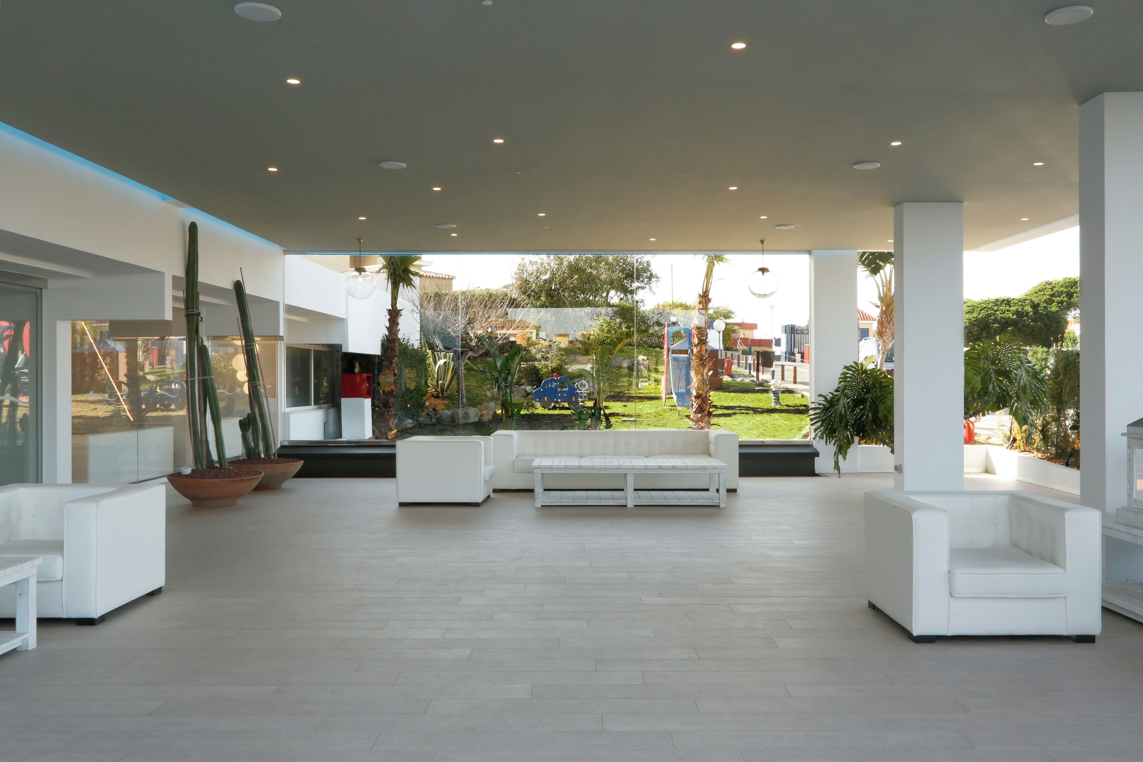 A spacious outdoor lounge area with white sofas and a coffee table, surrounded by potted plants and a view of a landscaped garden.