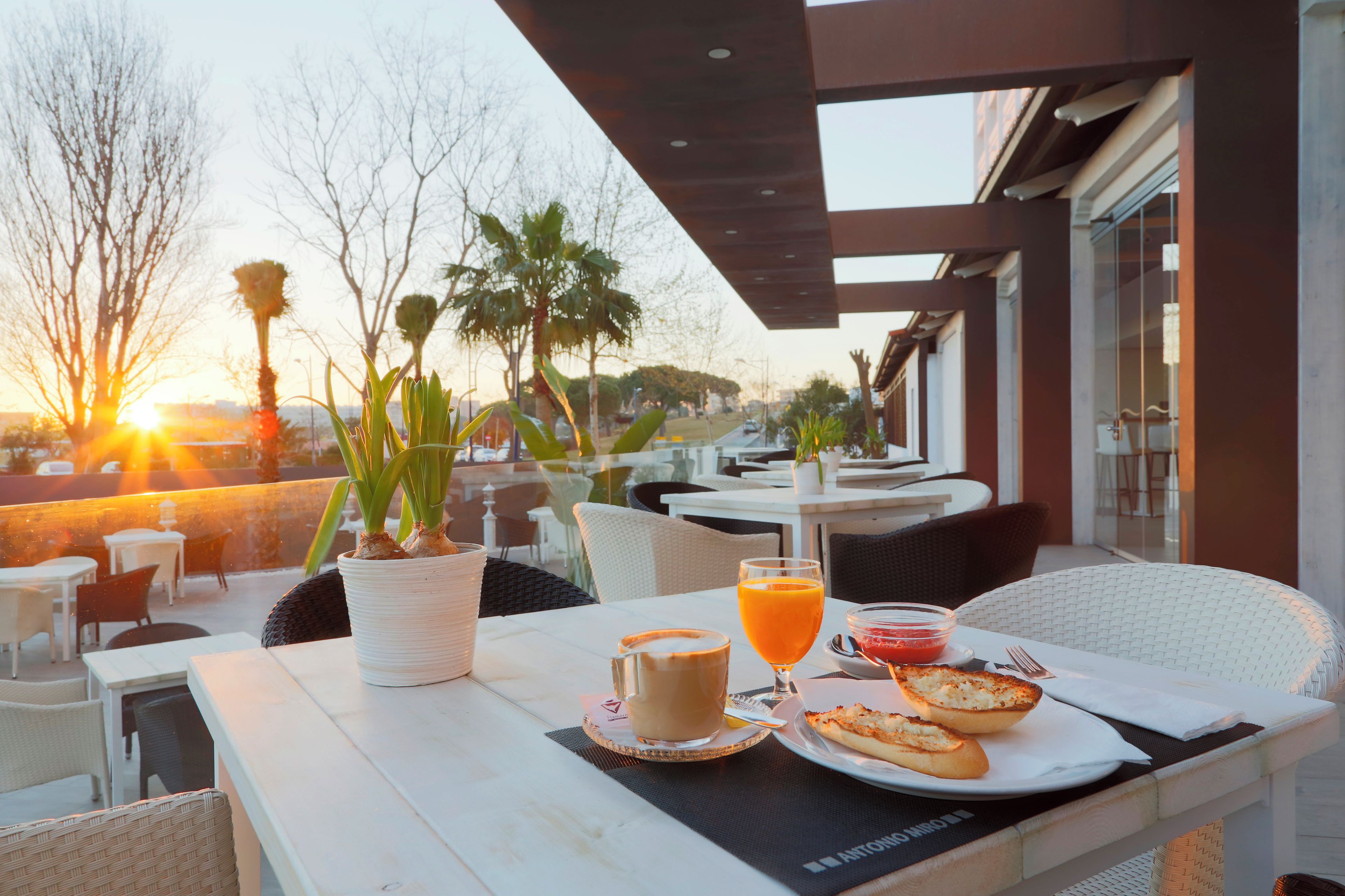 Un área de comedor al aire libre con una mesa dispuesta para dos personas, que incluye una bebida, un pastel y una planta.