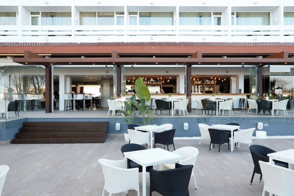 An outdoor seating area with tables and chairs, a bar in the background, and a building with multiple floors and balconies. - Flamero (Almonte)