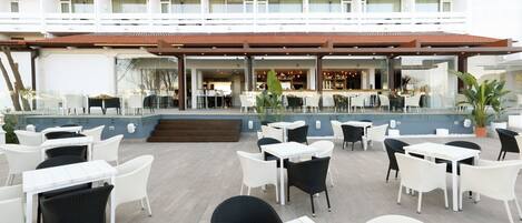 Un espace extérieur aménagé pour les repas, avec des chaises et des tables en osier blanches et noires, un coin bar doté d’un comptoir, et une piscine en arrière-plan.