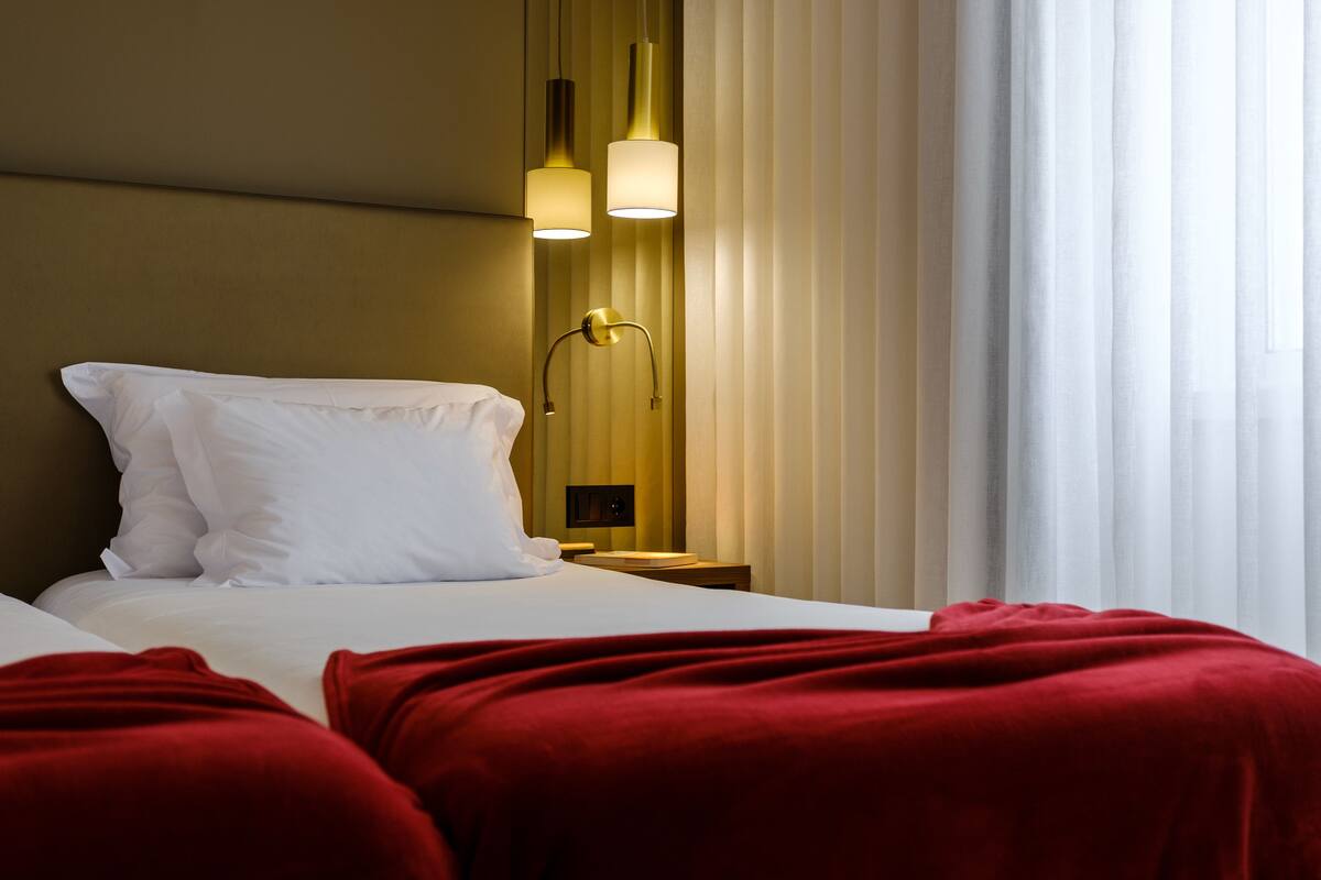A hotel room with a bed, a nightstand, a wall-mounted lamp, and a window with curtains.
