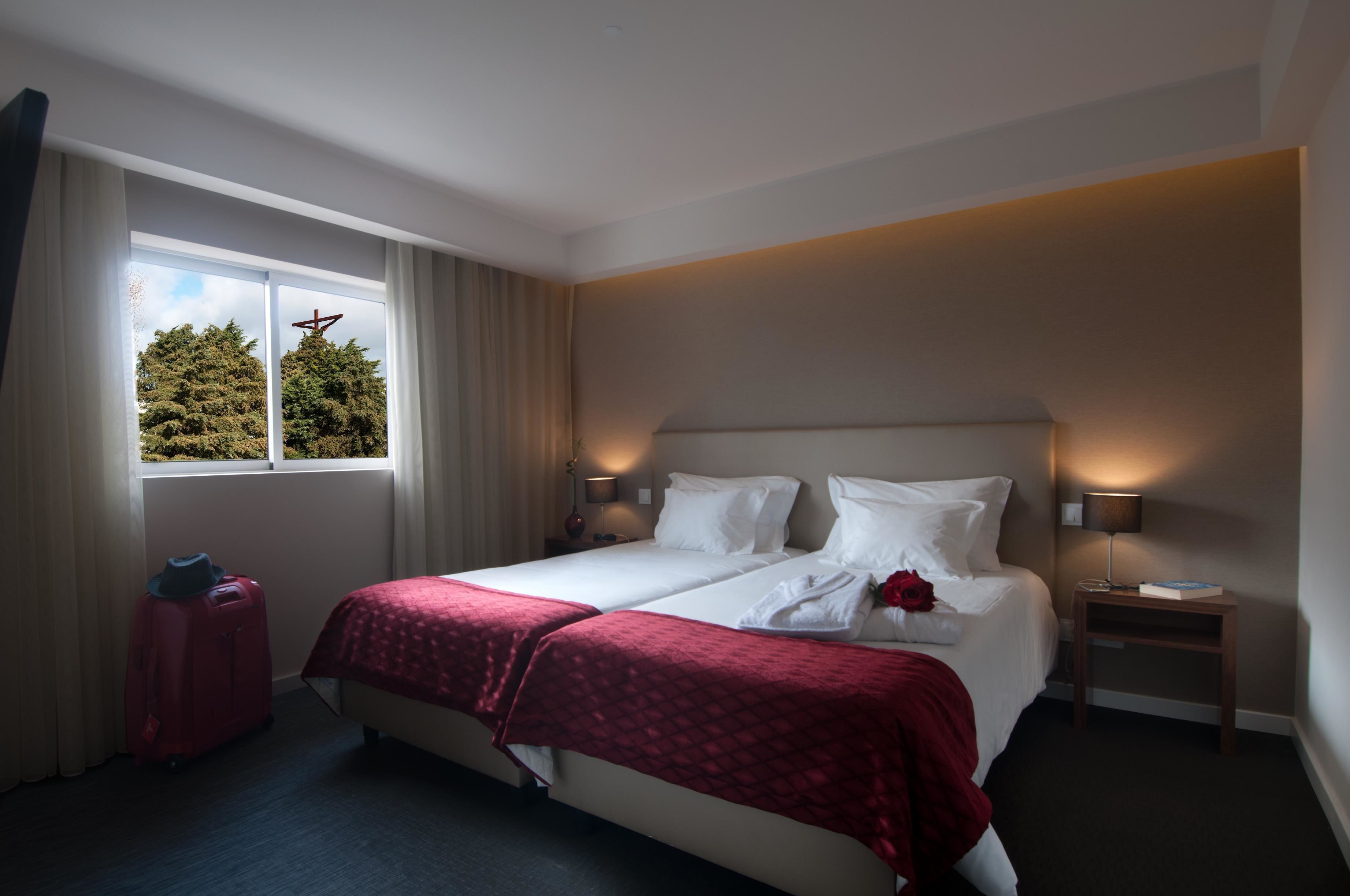 Une chambre d’hôtel avec un lit, deux lampes de chevet, une table de nuit, une fenêtre avec des rideaux et une valise rouge.