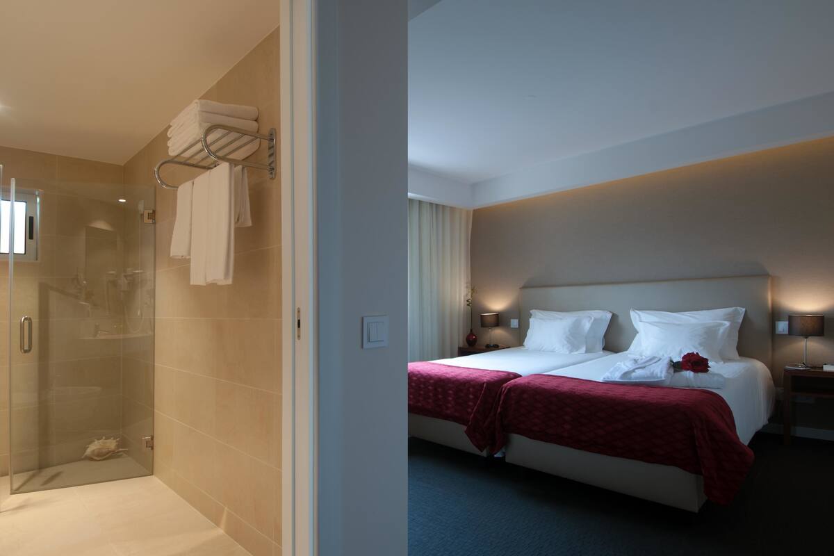 A hotel room with a glass-enclosed shower, a neatly made bed with a red blanket, and a bedside table with a lamp.
