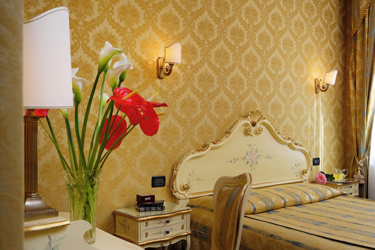 Una habitación clásica con cama, mesita de noche, lámpara y un jarrón con flores.