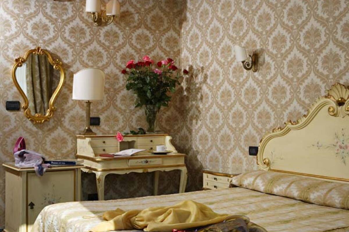 Een slaapkamer met een bed, een kaptafel met spiegel, een lamp en een vaas met bloemen.