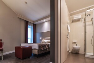 Una camera d'albergo con un letto, un poggiapiedi rosso, una finestra con tende e un box doccia con porta in vetro.