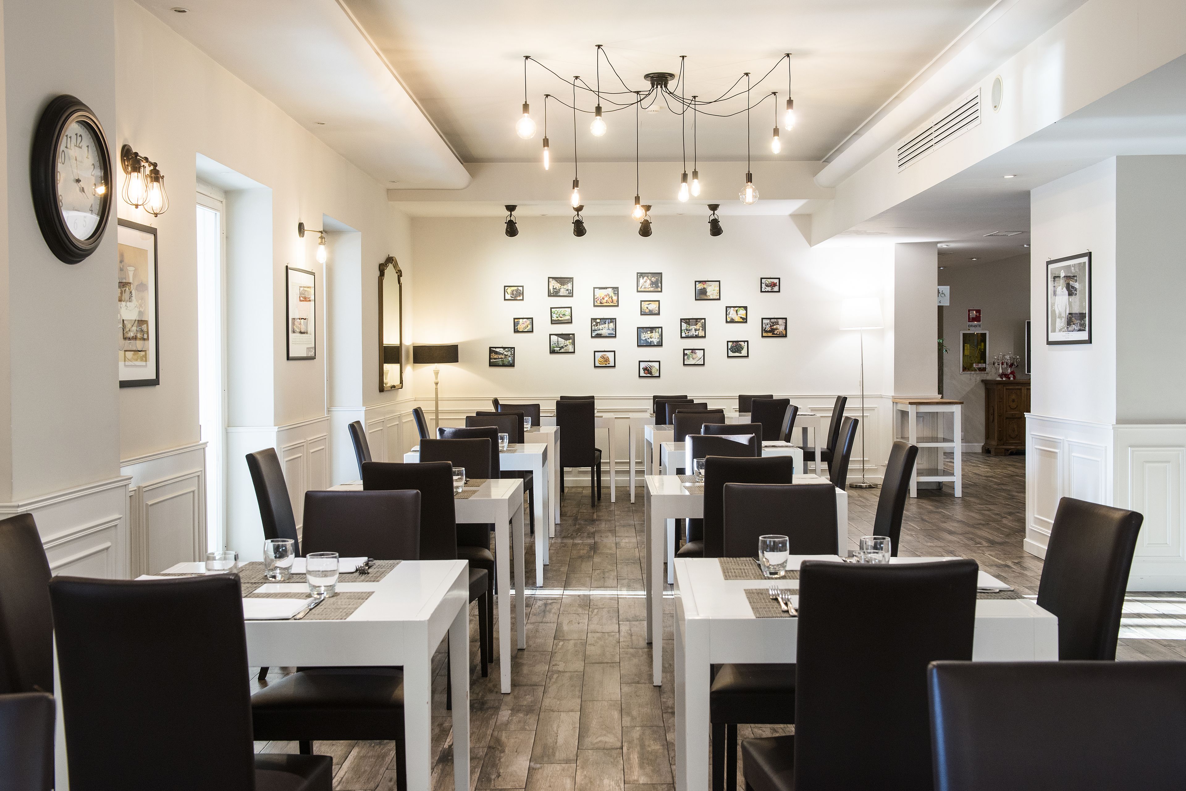 A dining area with multiple tables and chairs, a chandelier, framed pictures on the wall, and a clock on the wall.