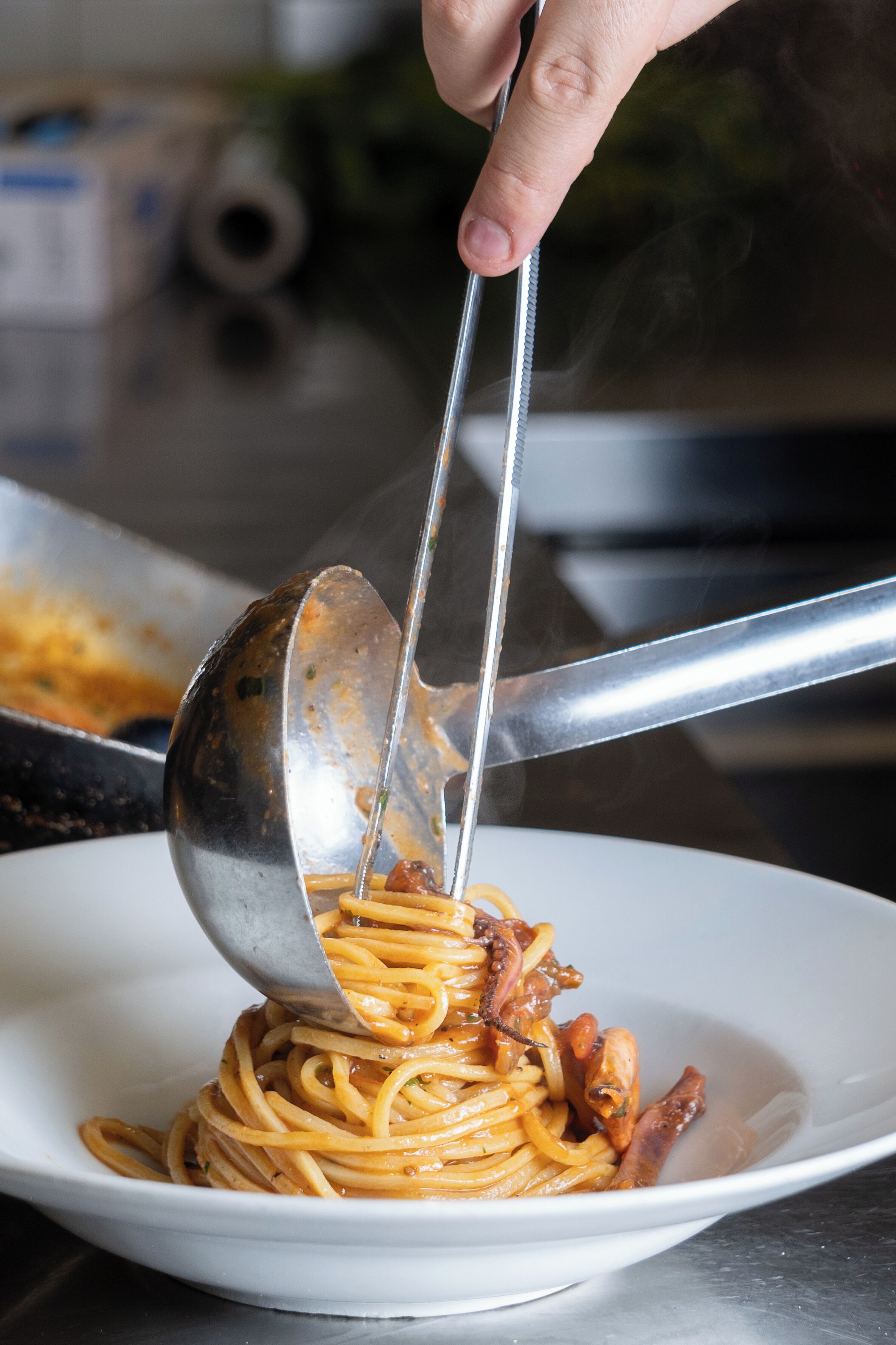 A hand using tongs to serve spaghetti onto a plate.