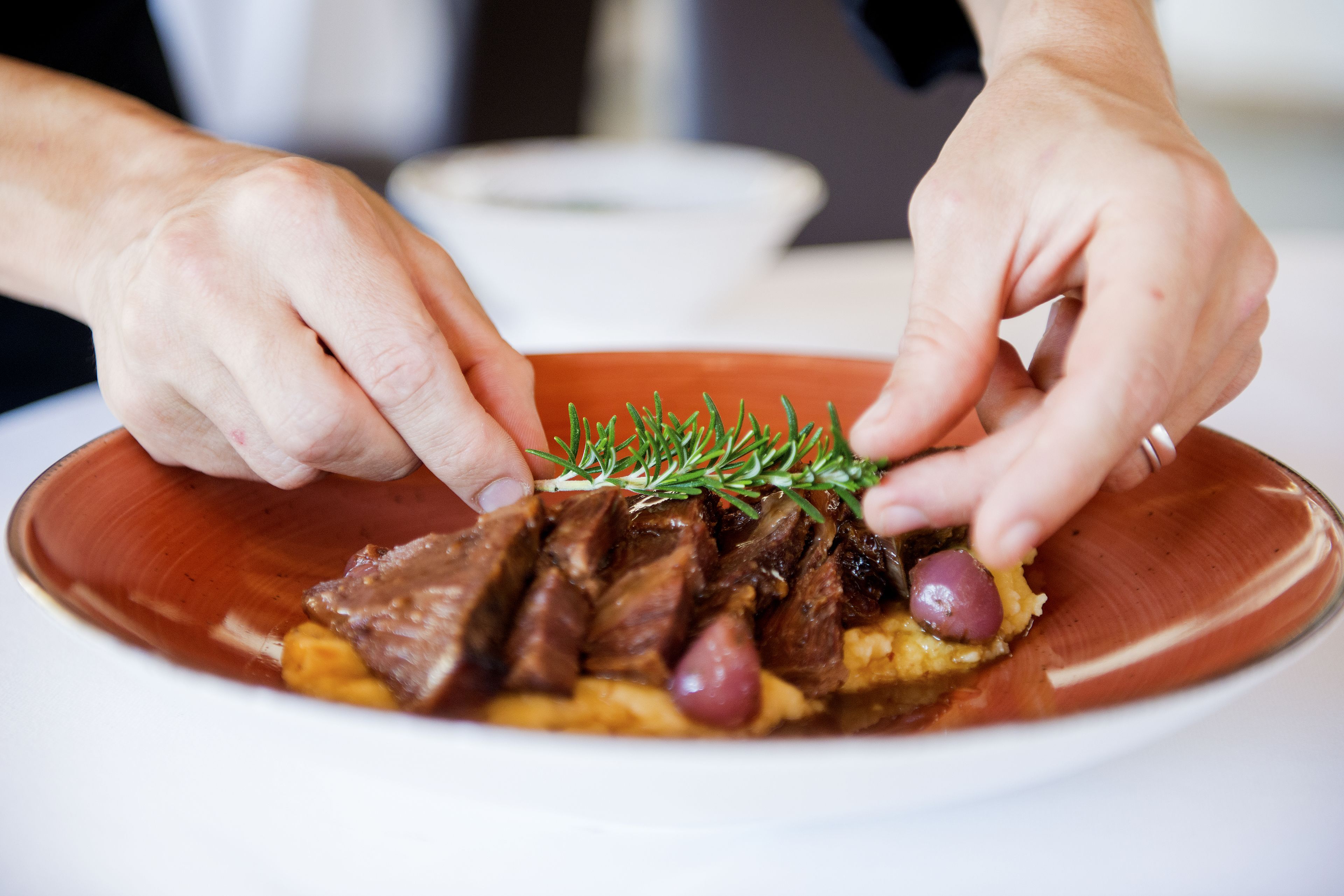 Une personne dépose une branche de romarin sur un plat composé de tranches de viande et de pommes de terre.