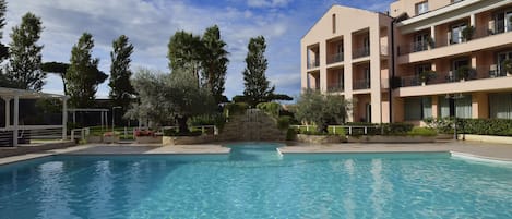 A clear blue pool surrounded by a multi-story building with balconies, trees, and a stone structure in the background.