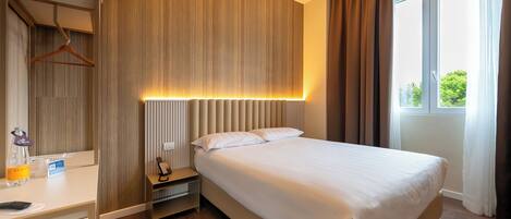 A hotel room with a bed, bedside table, and a window with curtains.