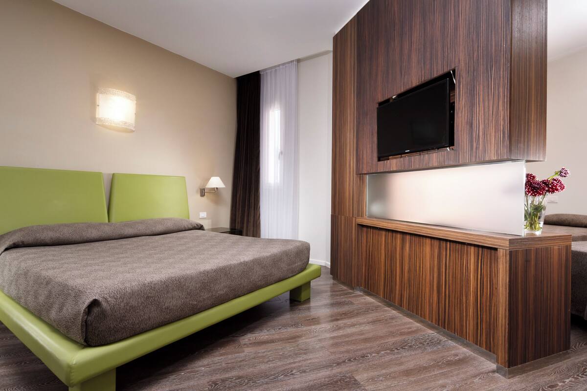 Una habitación de hotel moderna con una cama verde, un panel de pared de madera, un televisor de pantalla plana y un florero con flores.