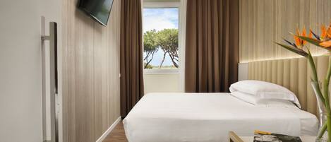 A hotel room with a bed, a TV mounted on the wall, a window with curtains, and a plant on the bedside table.