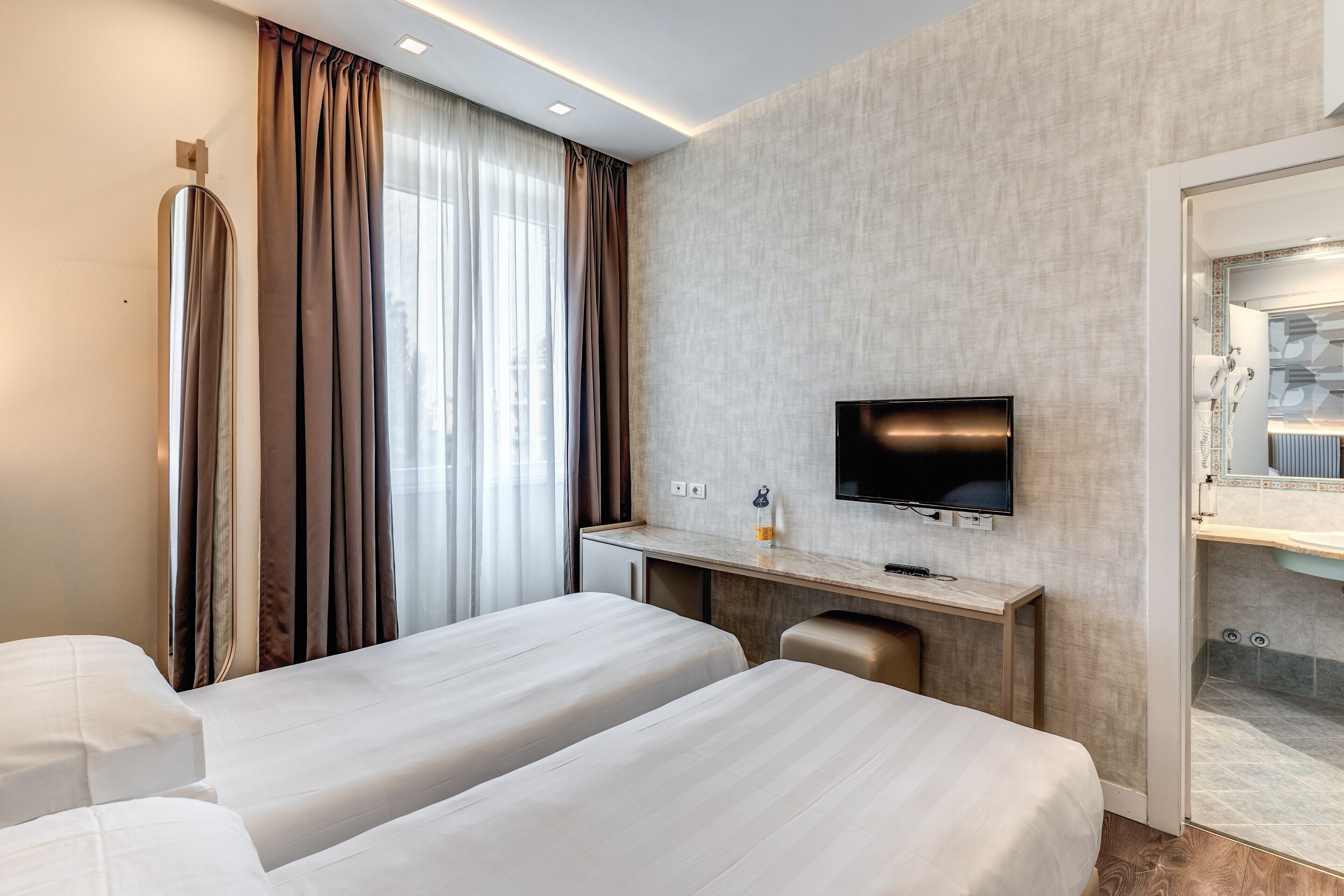 Quarto de hotel com duas camas, uma mesa de escritório com televisão de tela plana, banheiro com espelho e porta-toalhas, e janela com cortinas.