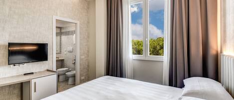 A modern hotel room with a large bed, a TV, a bathroom, and a window with curtains.