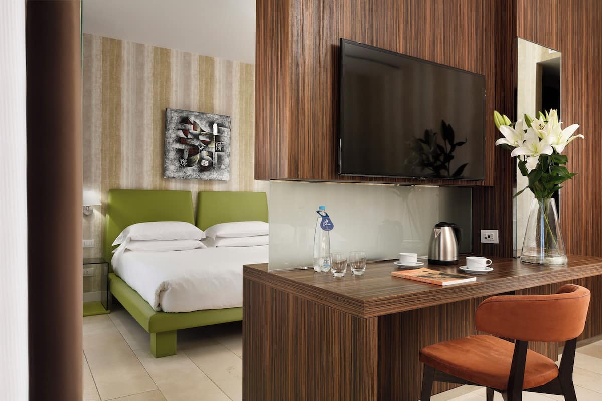 Una habitación de hotel con una cama, un escritorio con una silla, un televisor, un espejo, un jarrón con flores y una cafetera.